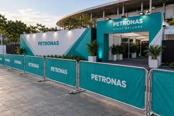 PETRONAS branded barricades and sponsor boundary with a gently angled row of realistic interlocking steel crowd-control barricades, each with breathable teal printed mesh covers showing simple PETRONAS text, exposed steel feet and frame edges, a separate matching sponsor media wall and event entrance behind